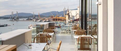 A terrace with tables and chairs overlooking a marina with boats and cranes.