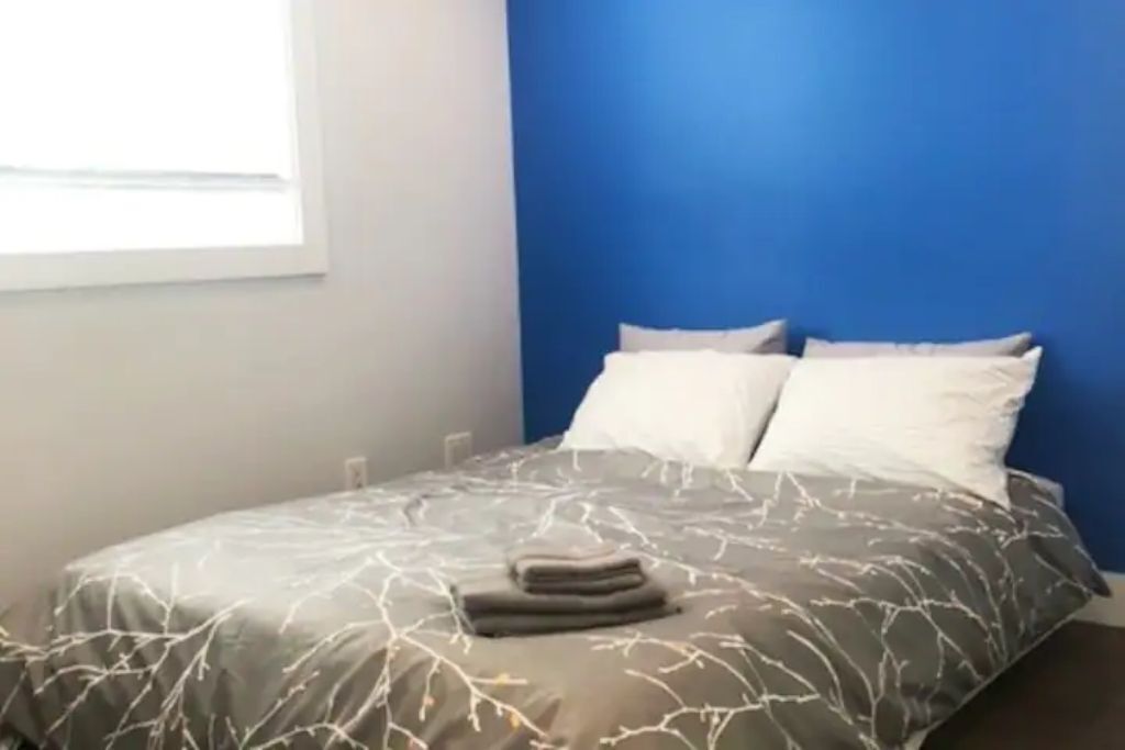 A bedroom with a bed against a blue wall, a window on the left, and a white pillow on the bed.