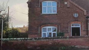 Une maison à deux étages, construite en briques, avec un toit en pente, une porte d’entrée et une petite fenêtre ronde à l’étage supérieur.