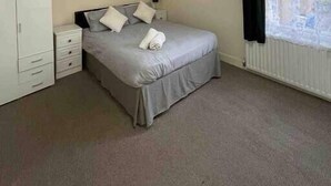 Un dormitorio con cama, armario, mesita de noche, un cuadro y ventana con cortinas.