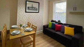 Un comedor con mesa de madera dispuesta para cuatro, un sofá negro con cojines de colores, un cuadro en la pared y una lámpara en el techo.
