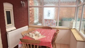 A dining area with a red polka dot tablecloth, wooden chairs, a white radiator, and a window with a view of a garden.