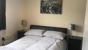 Un lit bien fait, avec une tête de lit, deux tables de chevet avec des lampes, un tableau accroché au mur et un luminaire au plafond.