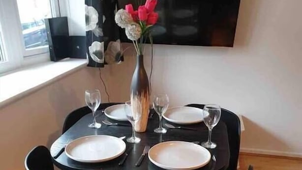 Une table à manger dressée pour quatre personnes, agrémentée d’un vase de fleurs, un téléviseur à écran plat fixé au mur et une fenêtre avec des rideaux.