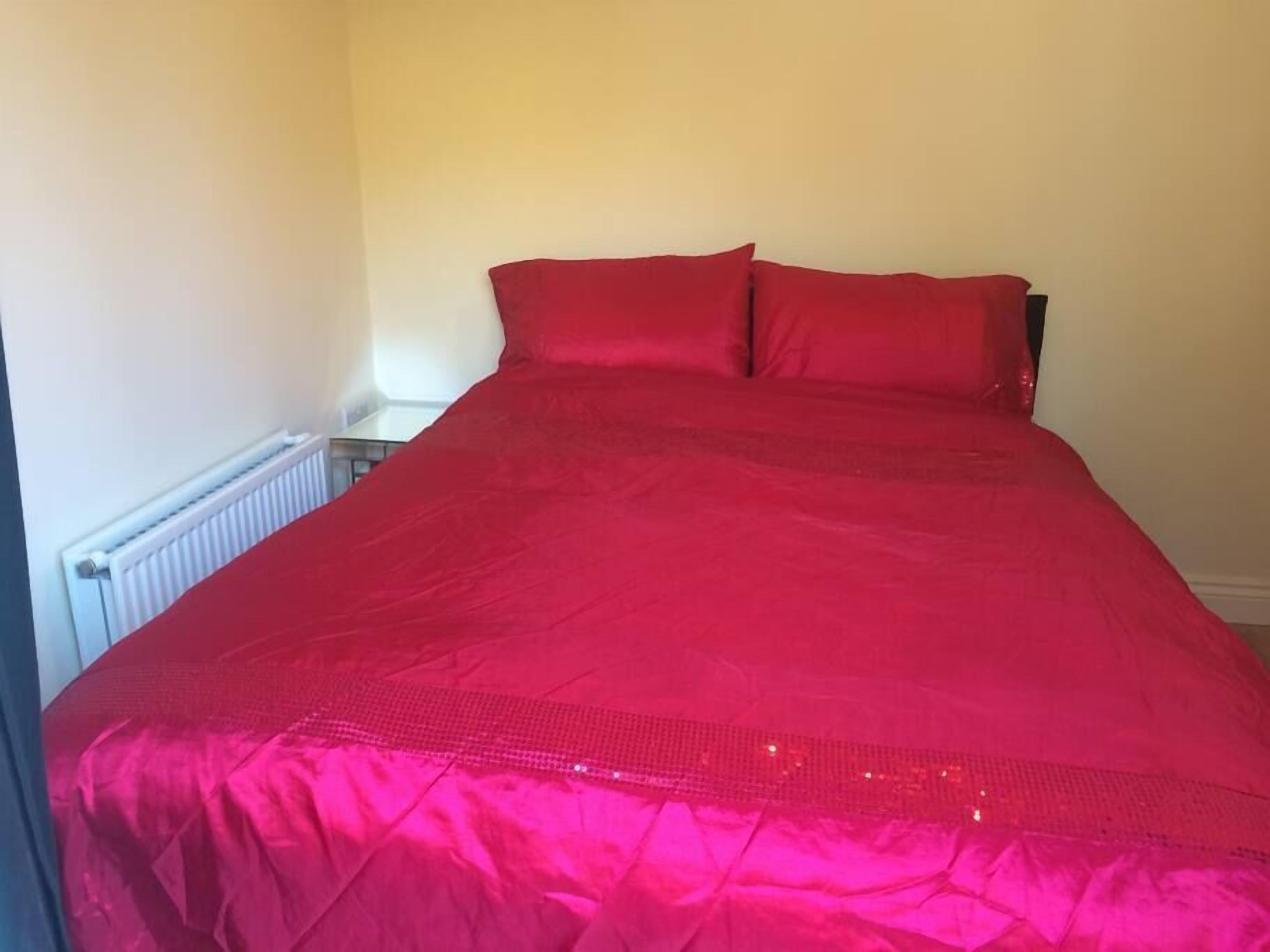 A bed with a red bedspread in a room with a radiator and a small table.