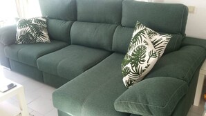Un canapé vert orné de coussins à motifs de feuilles, dans une pièce au sol blanc et à la table également blanche.