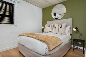 Une chambre à coucher avec un lit, un miroir rond, une fenêtre avec des stores et une table de chevet avec une lampe.