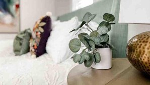 Ein ordentlich eingerichteter Raum mit einem Bett, einem weißen Kopfkissen, einer grünen Pflanze im weißen Topf und einem dekorativen Gegenstand auf dem Nachttisch.