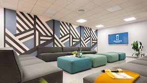 A modern living room with grey sofas, teal ottomans, and a large geometric wall design. - Micampus Rector Estanislao del Campo - Students Residence (Seville)