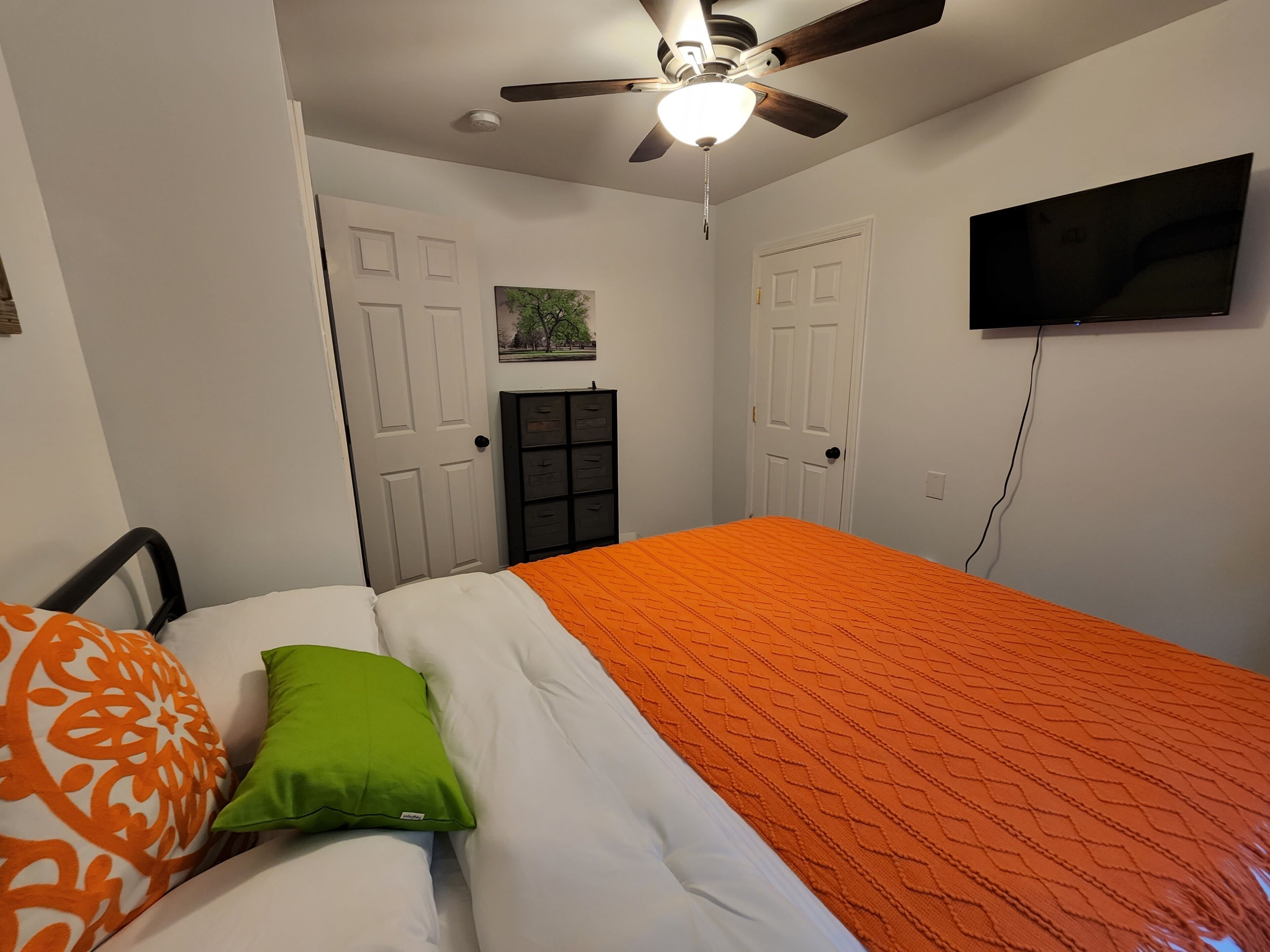 A bedroom with a bed, a ceiling fan, a TV mounted on the wall, and a door with a small window.