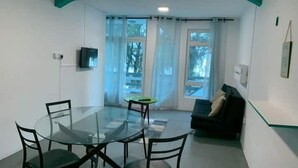 Een moderne woonkamer met een glazen eettafel, twee stoelen, een bank en een aan de muur bevestigde televisie.