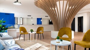 A modern lobby with a reception desk, yellow chairs, a white sofa, and a unique wooden ceiling feature. - Micampus Armendáriz - Students Residence (Seville)