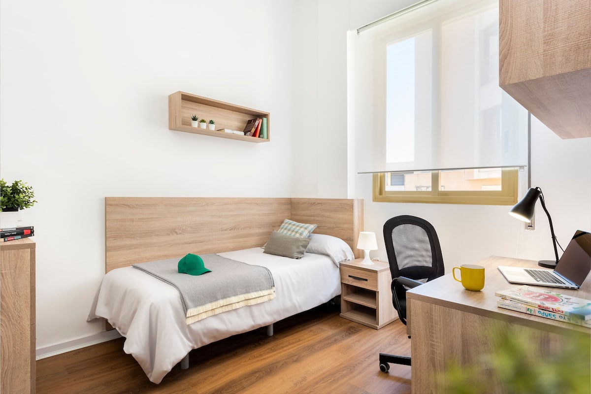 Ein ordentlich eingerichtetes Schlafzimmer mit Bett, Nachttisch, Sessel, Schreibtisch und Fenster.