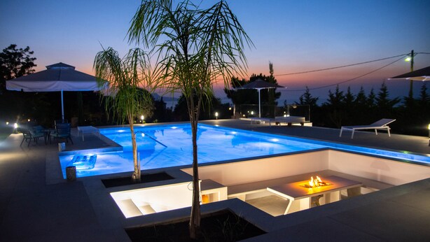 A swimming pool with a fire pit and lounge chairs, surrounded by umbrellas and a sunset view. - Villa Mediterranean Oasis (Rethymno)