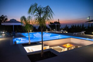 A swimming pool with a fire pit and lounge chairs, surrounded by umbrellas and a sunset view. - Villa Mediterranean Oasis (Rethymno)