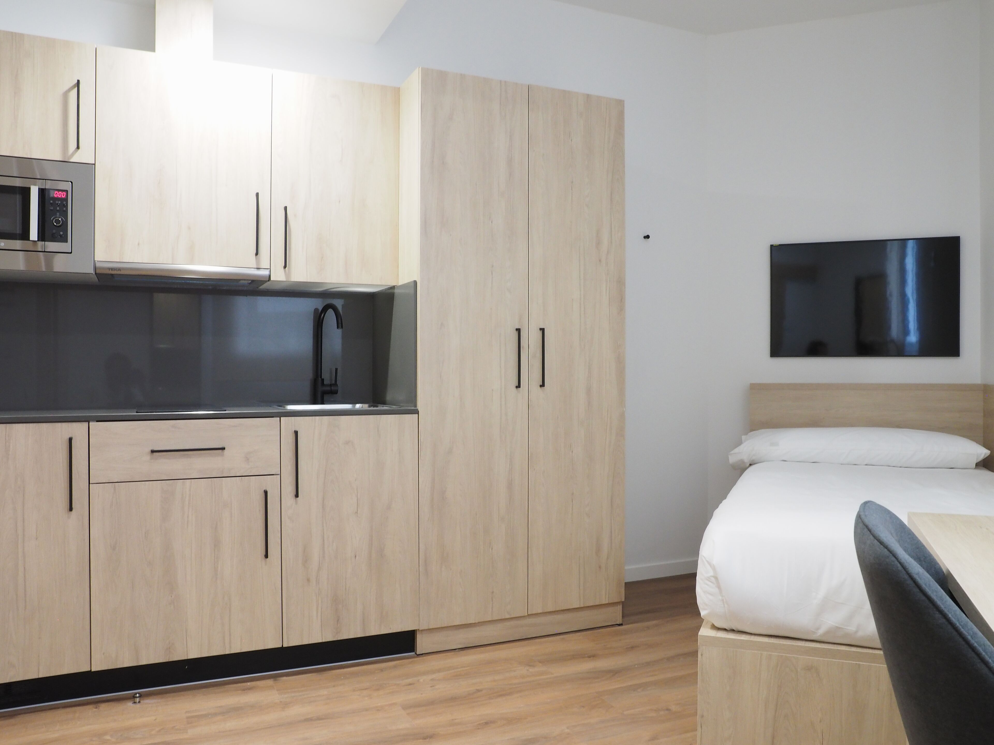 Ein kompaktes Hotelzimmer mit Küchenzeile, einem Bett mit weißer Bettwäsche und einem wandmontierten Flachbildfernseher.