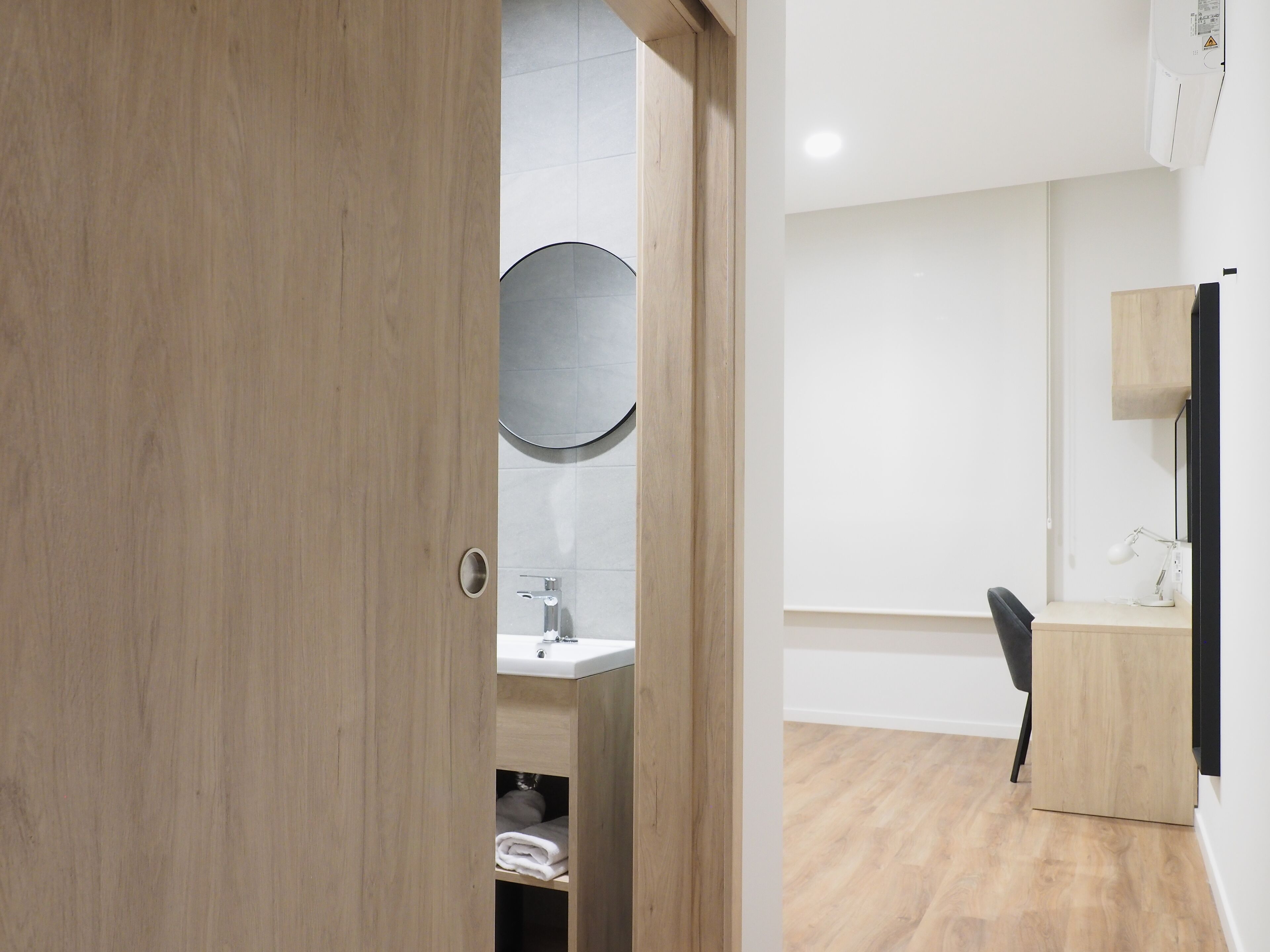 A modern bathroom with a wooden door, a round mirror, a sink, and a black chair.