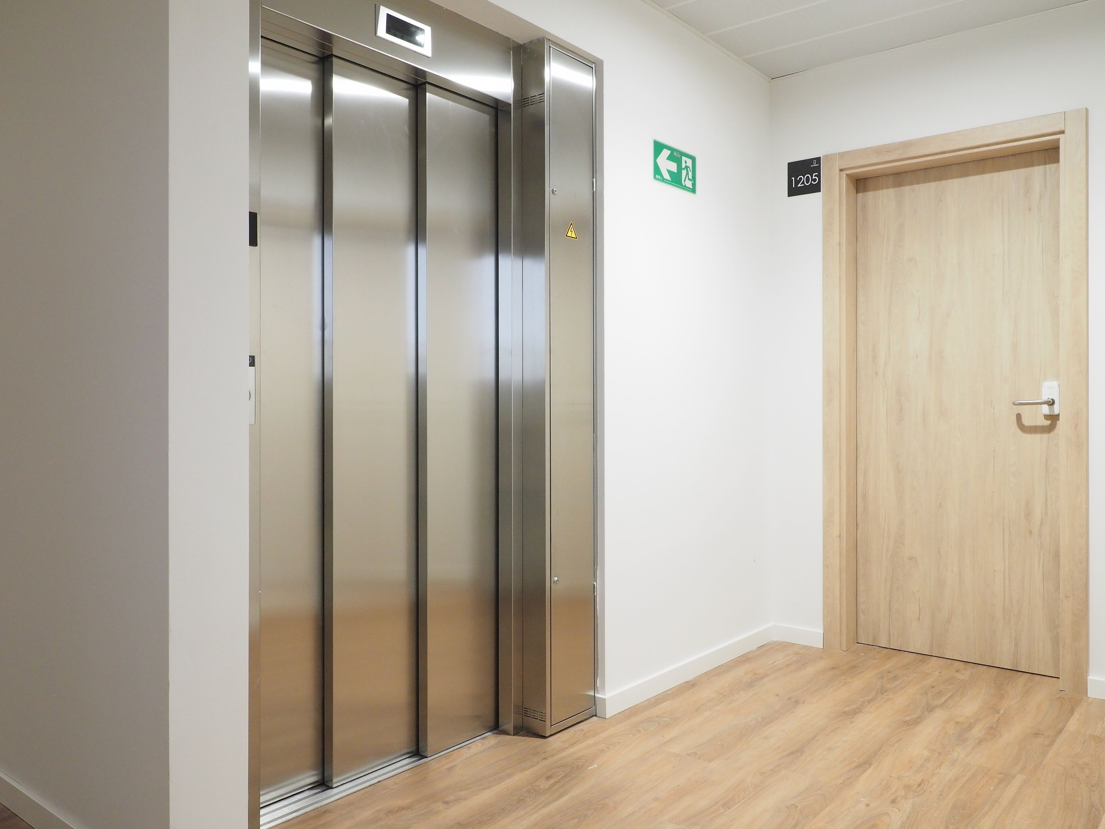 Un ascensor moderno con detalles metálicos, una puerta de madera a la derecha y un letrero de salida de emergencia verde en la pared.