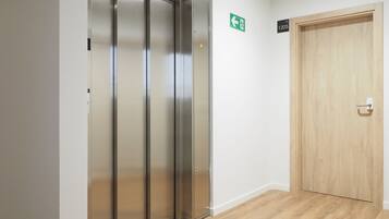 Un ascensor moderno con detalles metálicos, una puerta de madera a la derecha y un letrero de salida de emergencia verde en la pared.