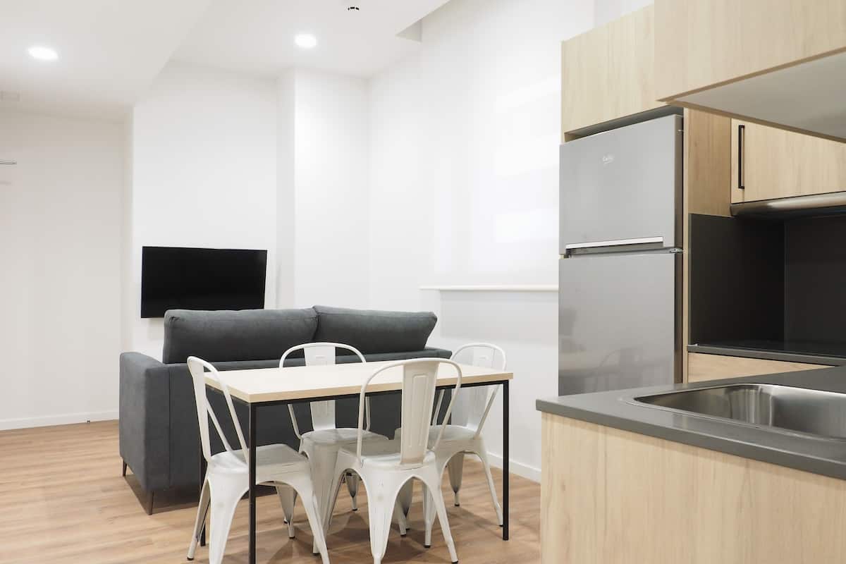A modern kitchen with a dining area, including a grey sofa, white chairs, and a flat-screen TV mounted on the wall.