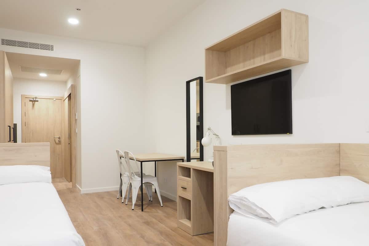 A modern hotel room with two beds, a desk with a chair, a TV, and a wooden shelf.