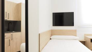 Habitación de hotel compacta con cama, mesita de noche pequeña, lavamanos integrado y televisor de pantalla plana montado en la pared.