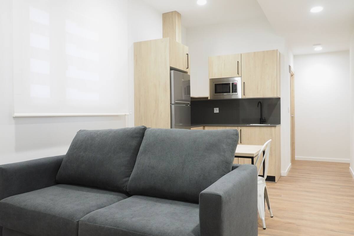 A modern living room with a grey sofa, a small dining table, and a kitchen area with wooden cabinets and stainless steel appliances.
