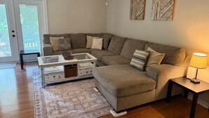 A living room with a sectional sofa, a glass coffee table, a side table with a lamp, and two wall art pieces.
