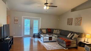 A living room with a grey sofa, a coffee table, a TV stand, and a ceiling fan. There is a sliding glass door leading to an outdoor area.