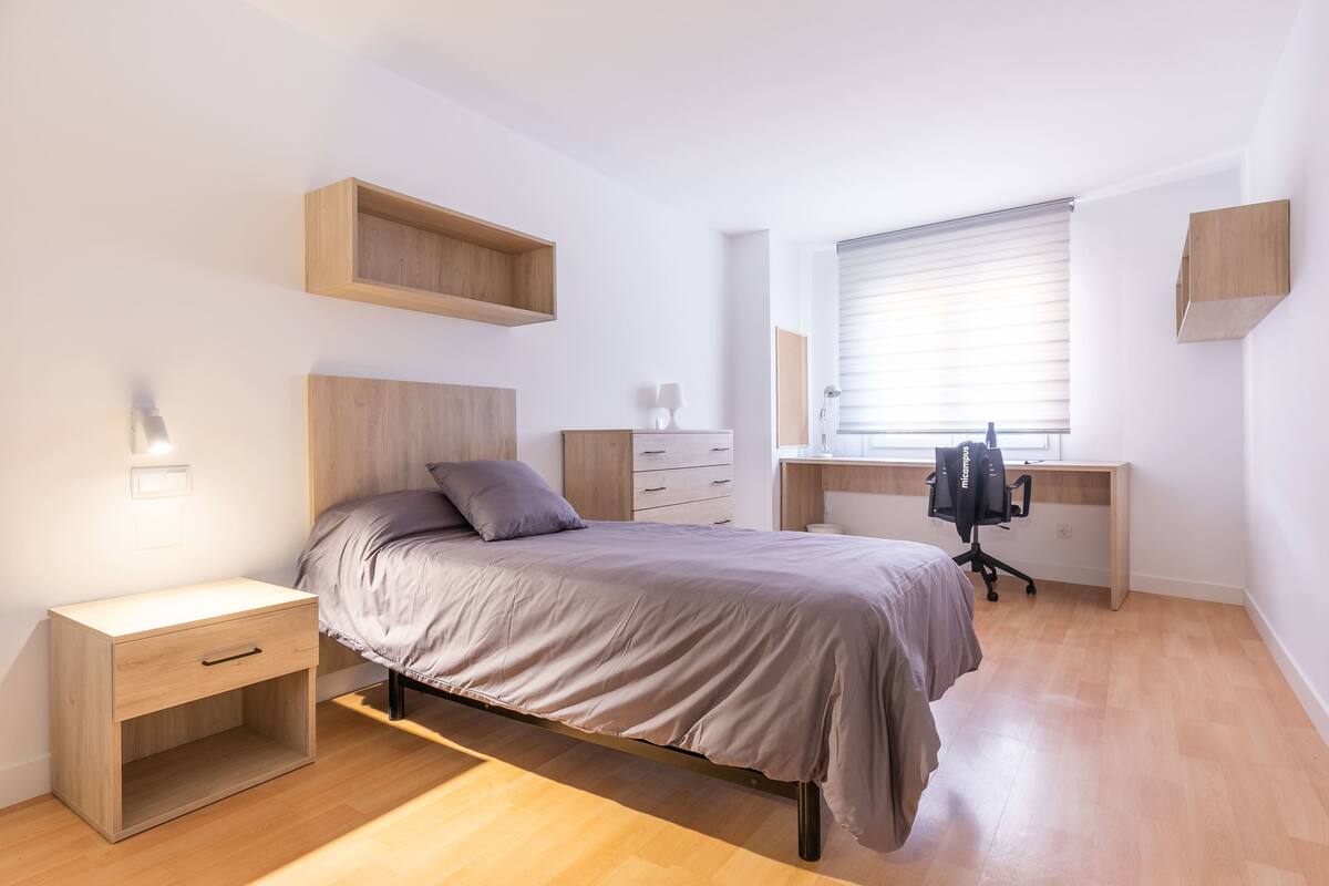 Un dormitorio con cama, escritorio y silla. Se ve una ventana con persianas, una mesita de noche y una lámpara.