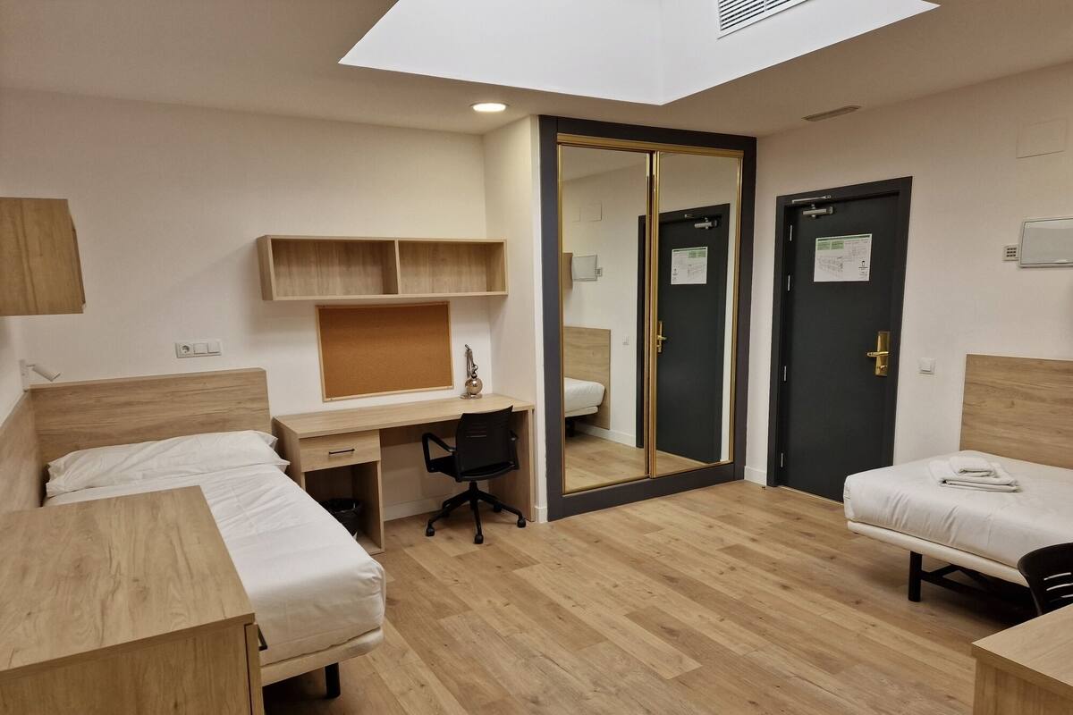 Una habitación de hotel compacta con dos camas individuales, un escritorio con silla, un armario pequeño y un espejo.