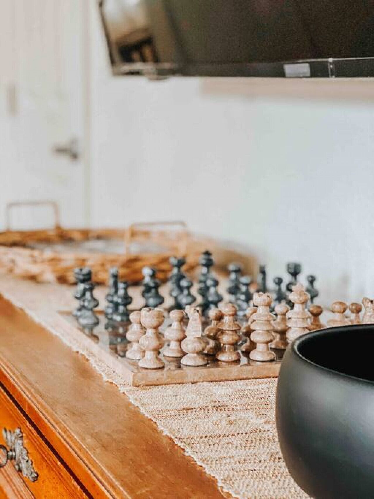 A chessboard on a wooden table with a black bowl beside it, in a room with a television mounted on the wall.