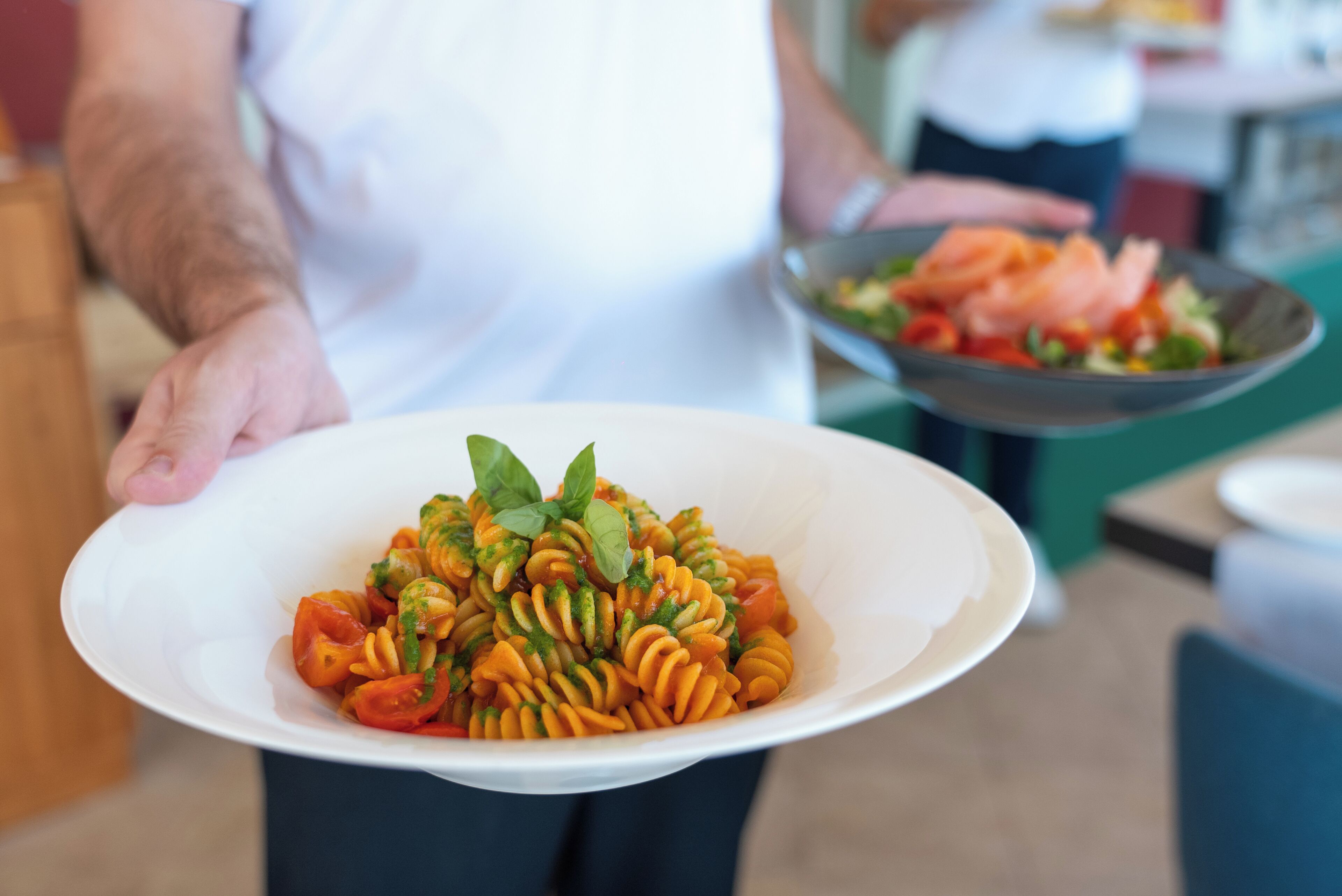 A person holding a white bowl with a pasta dish garnished with basil.