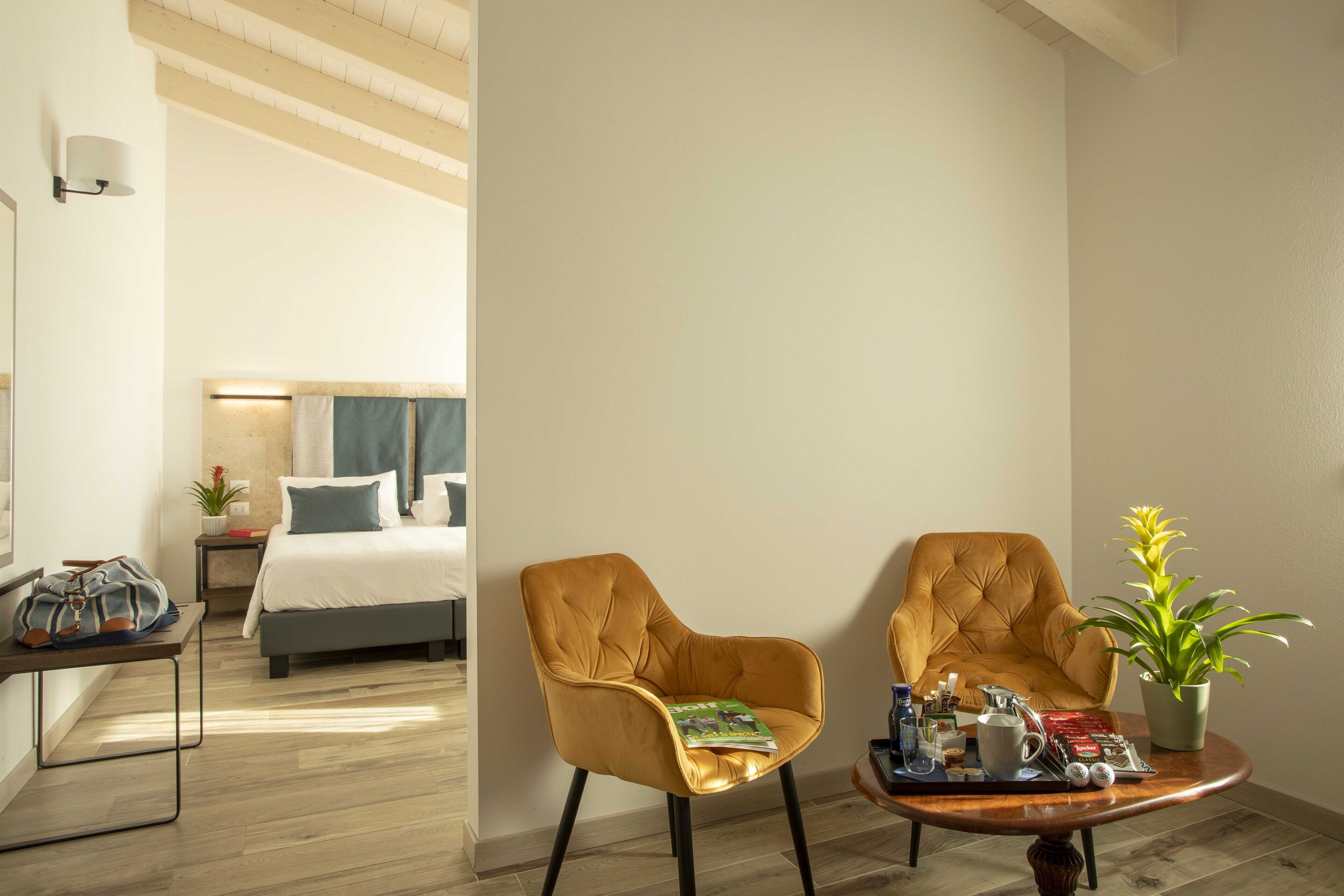 A hotel room with a bed, two armchairs, a coffee table with a magazine and coffee items, a potted plant, and a suitcase on a side table.
