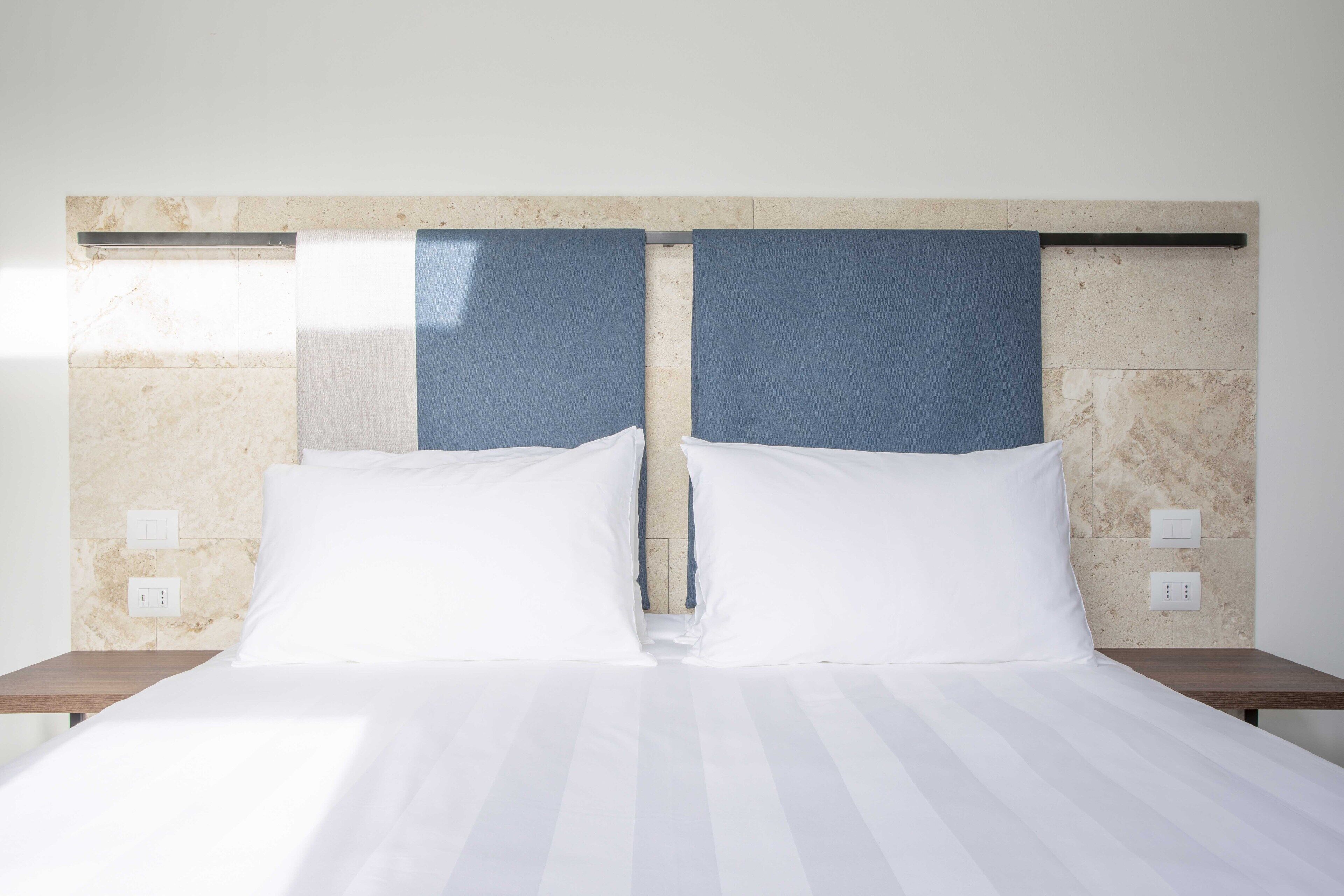 Una cama bien hecha con sábanas blancas y dos almohadas, un cabecero con detalles azules y una pared con diseño de mármol.