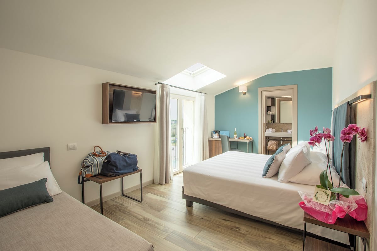 A hotel room with a bed, a desk, a chair, a table with a bag and a vase of flowers, and a window with curtains.