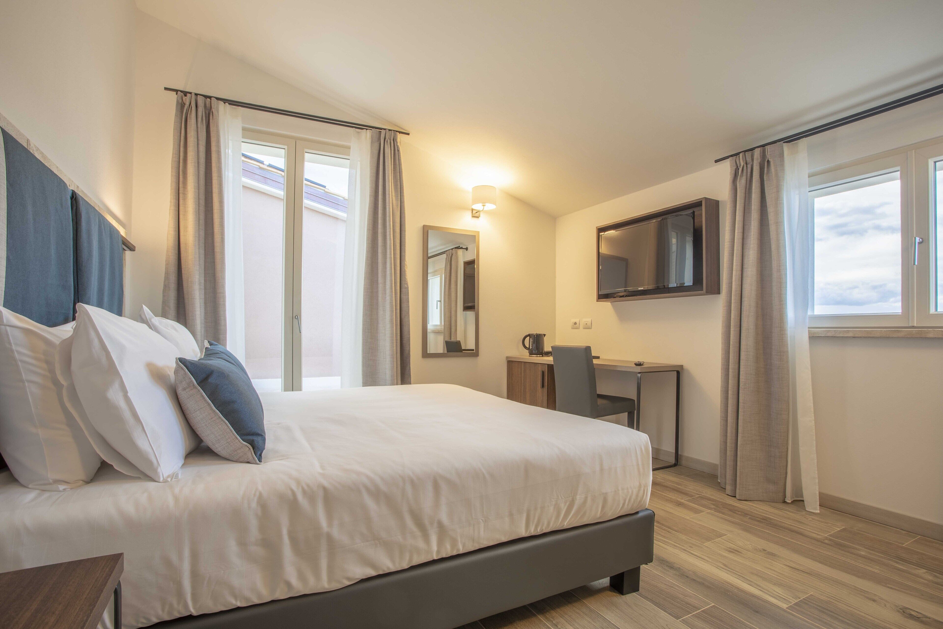 Una habitación de hotel moderna con una cama grande, un escritorio, una silla, un televisor de pantalla plana y un ventanal con cortinas.