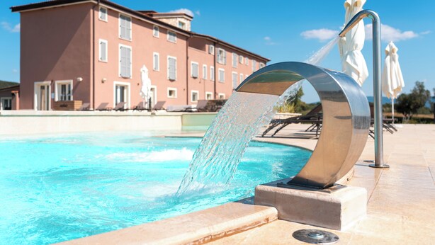A swimming pool with a water feature in the foreground and a building in the background. - Riva Toscana Golf Resort & SPA (Follonica)