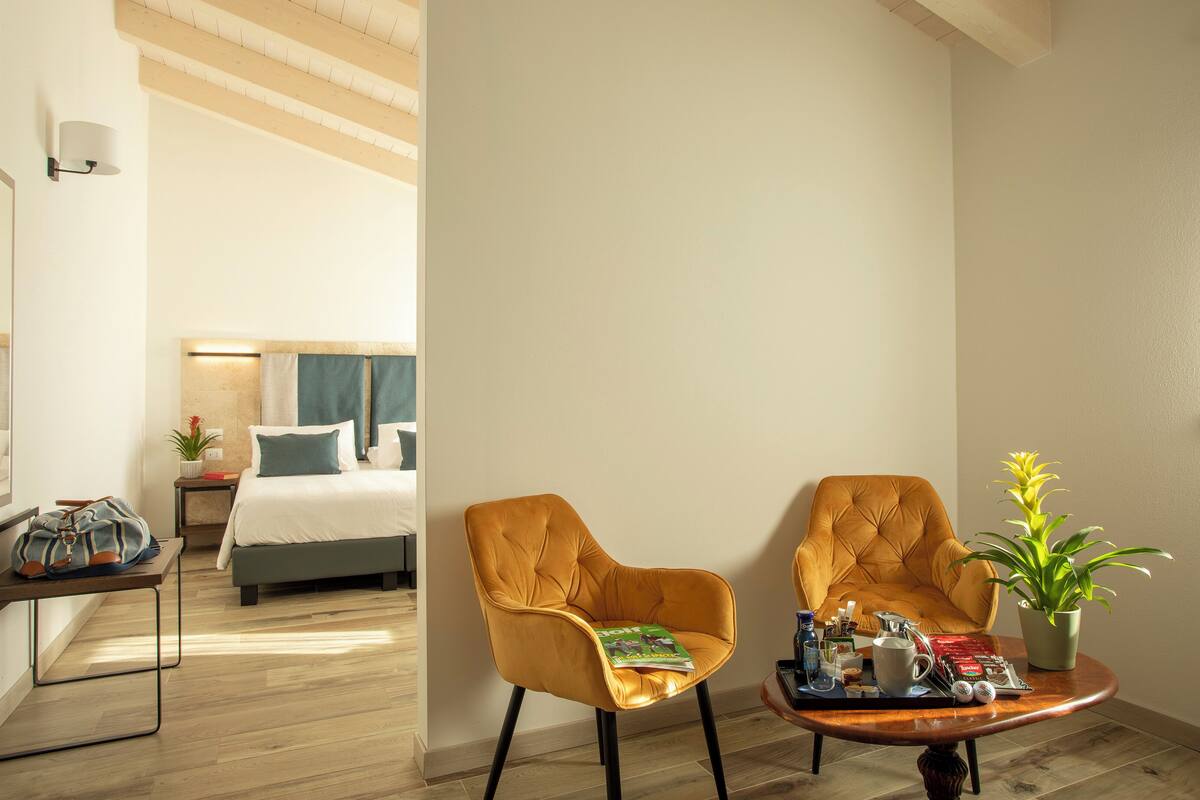 A hotel room with a bed, two armchairs, a coffee table with a magazine, a plant, and a lamp.