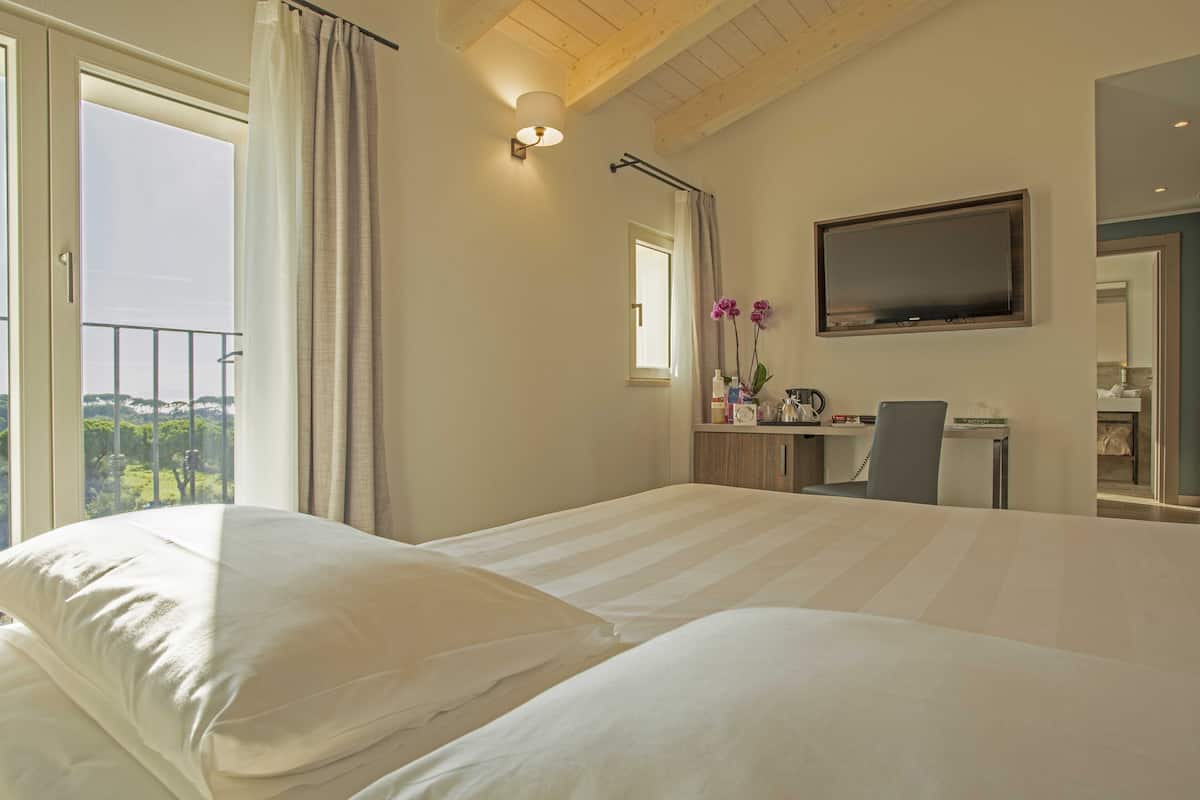 A hotel room with a large bed, a desk with a chair, a flat-screen TV, and a view of greenery through the window.