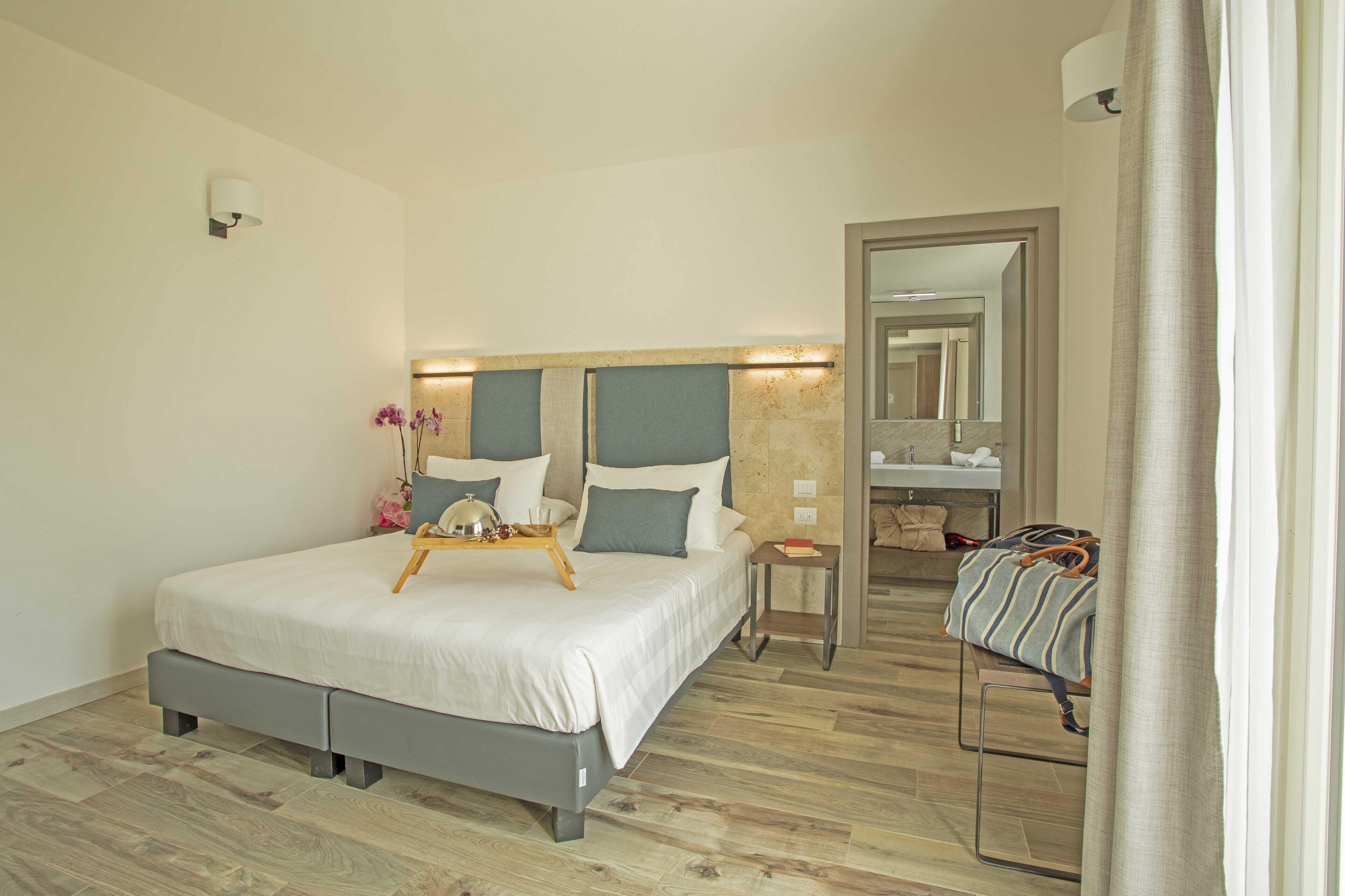 Quarto de hotel moderno com uma cama grande, mesas de cabeceira, um espelho e um banheiro visível através de uma porta aberta.