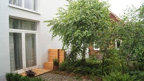 Un bâtiment moderne à l’extérieur blanc, avec un banc en bois et un petit espace jardin agrémenté de diverses plantes et d’un arbre.
