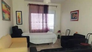 A room with a bed, a yellow armchair, a black sofa, and a small table with a lamp.