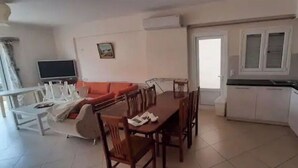 A dining area with a wooden table and chairs, a sofa, and a TV. There is a kitchen with white cabinets and a black countertop. - Family apartment 5 by Hillside apartments TM (Sarande)