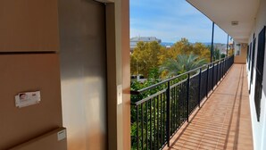 Un pasillo con una puerta, un balcón con barandilla y vistas a árboles y edificios.