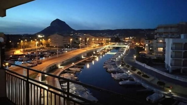Una vista nocturna de un puerto deportivo con barcos, un puente y edificios al fondo.