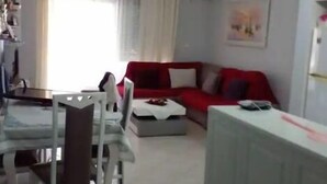 Uma sala de estar com um sofá vermelho, uma mesa de jantar com cadeiras, e uma cozinha ao fundo.