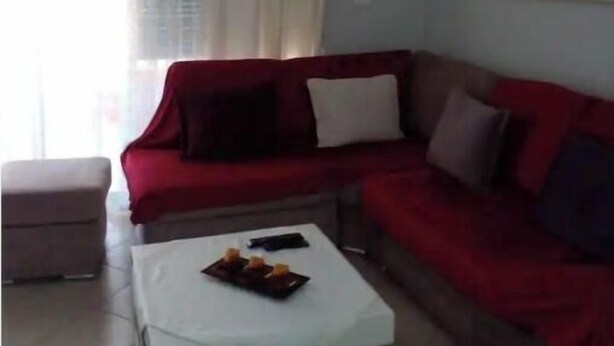 Uma sala de estar com um sofá vermelho, uma mesa de centro branca e um pequeno banquinho. Há uma janela com cortinas, um aparelho de ar condicionado na parede e um quadro sobre o sofá.