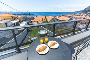 A balcony with a table set for breakfast, overlooking a coastal town. - Casa da Av by Atlantic Holiday (Machico)