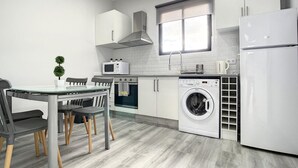 Een moderne keuken met witte kasten, een inbouwoven, magnetron en wasmachine. Er staat een eettafel met stoelen en een kleine plant in een pot.